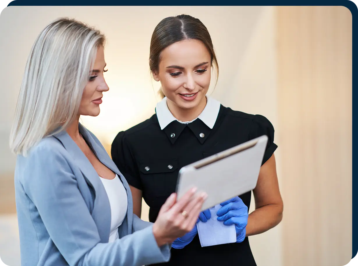 Two women looking at a tablet. One wears a black dress with gloves, and the other wears a light gray blazer.